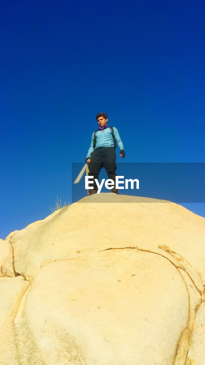 Low angle view of man holding knife while standing on rock against clear blue sky