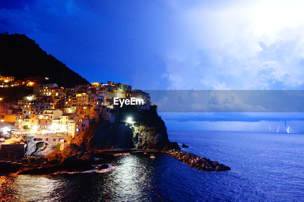 Illuminated buildings by sea against blue sky