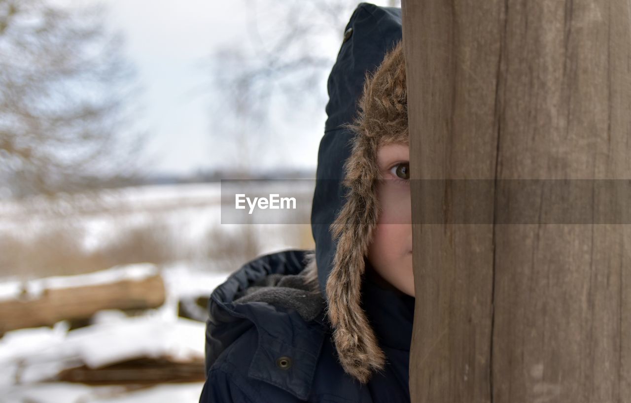 Close-up of boy wearing fur hat standing behind wooden fence during winter