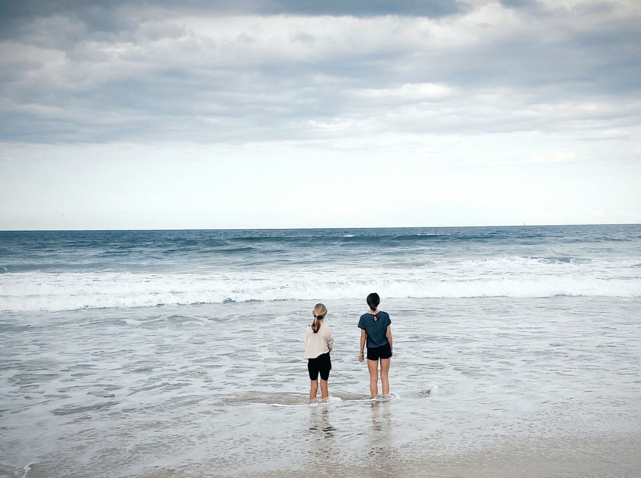 Rear view of girls standing in water at beach against cloudy sky