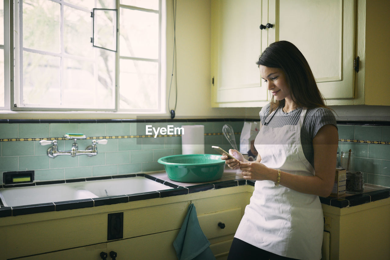 Young woman using mobile phone while standing at kitchen counter by window