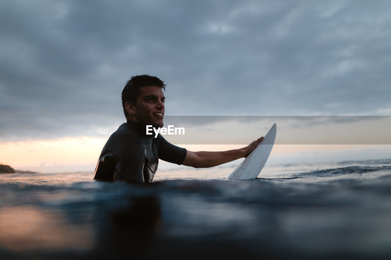 Cheerful male surfer on surfboard in wavy sea looking forward against mount in daytime