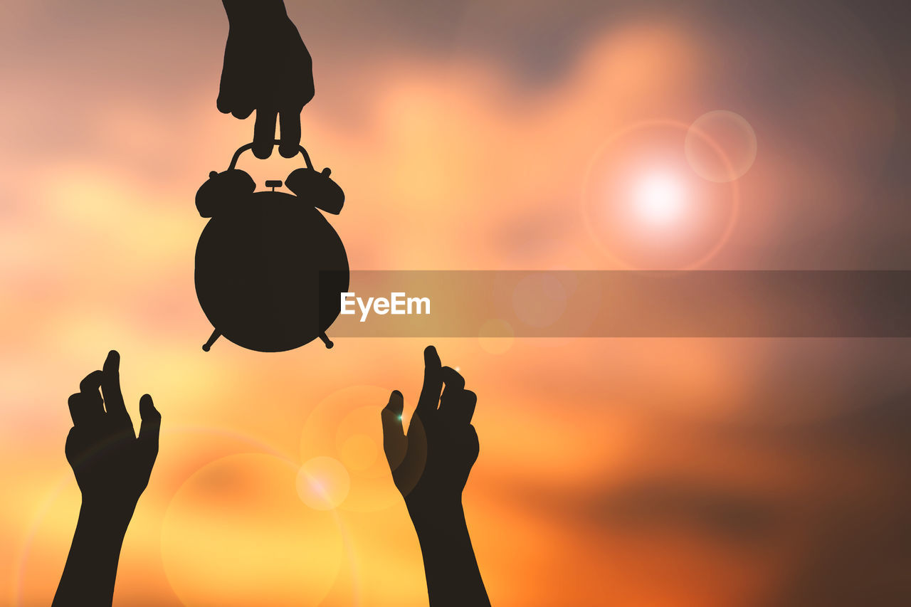 Cropped image of silhouette hands with alarm clock against sky during sunset