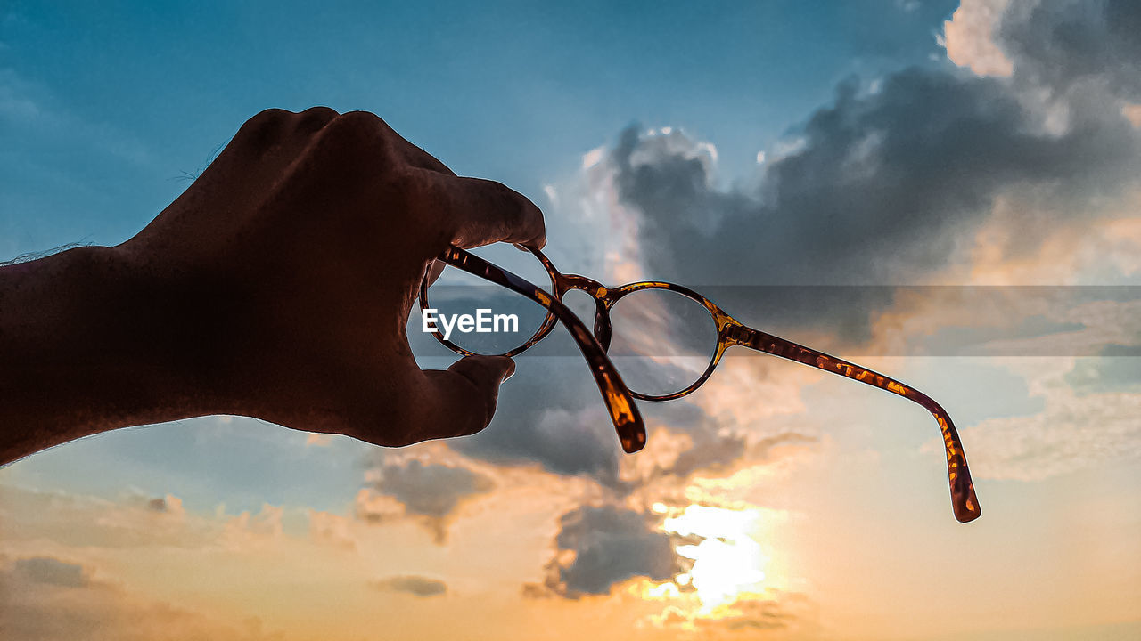 Close-up of hand holding eyeglasses against sky during sunset