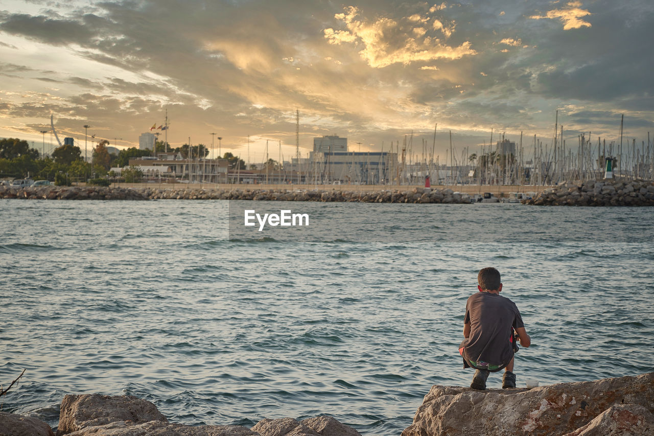 Rear view of man standing by sea against sky during sunset