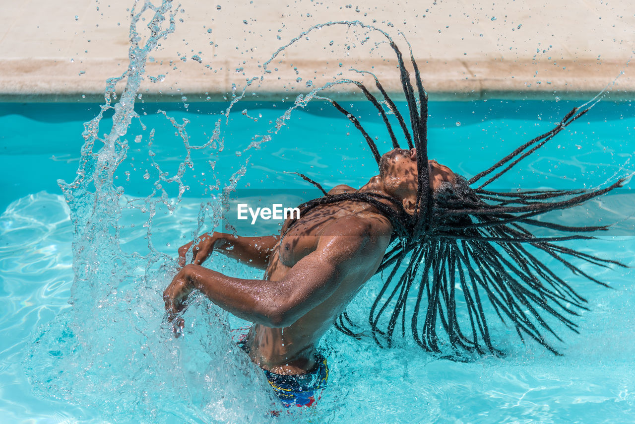 Black man with dreadlocks inside a pool raising his wet hair making a trail of water