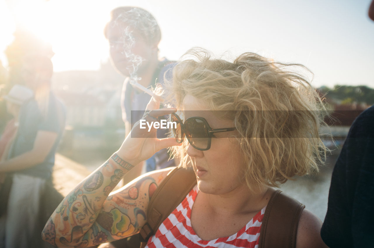 Woman wearing sunglasses while smoking cigarette in city at sunset