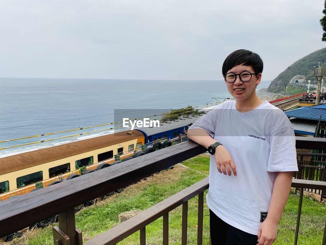 vacation, one person, water, smiling, glasses, railing, sea, casual clothing, standing, nature, young adult, adult, happiness, sky, lifestyles, portrait, leisure activity, vehicle, looking at camera, day, front view, eyeglasses, architecture, men, three quarter length, emotion, travel, transportation, outdoors, tourism, clothing, waist up, person, sunglasses, fashion, land, t-shirt, cheerful, holiday, beauty in nature, built structure, scenics - nature