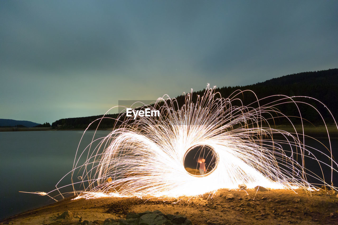 Man spinning illuminated wire wool by lake against sky at night