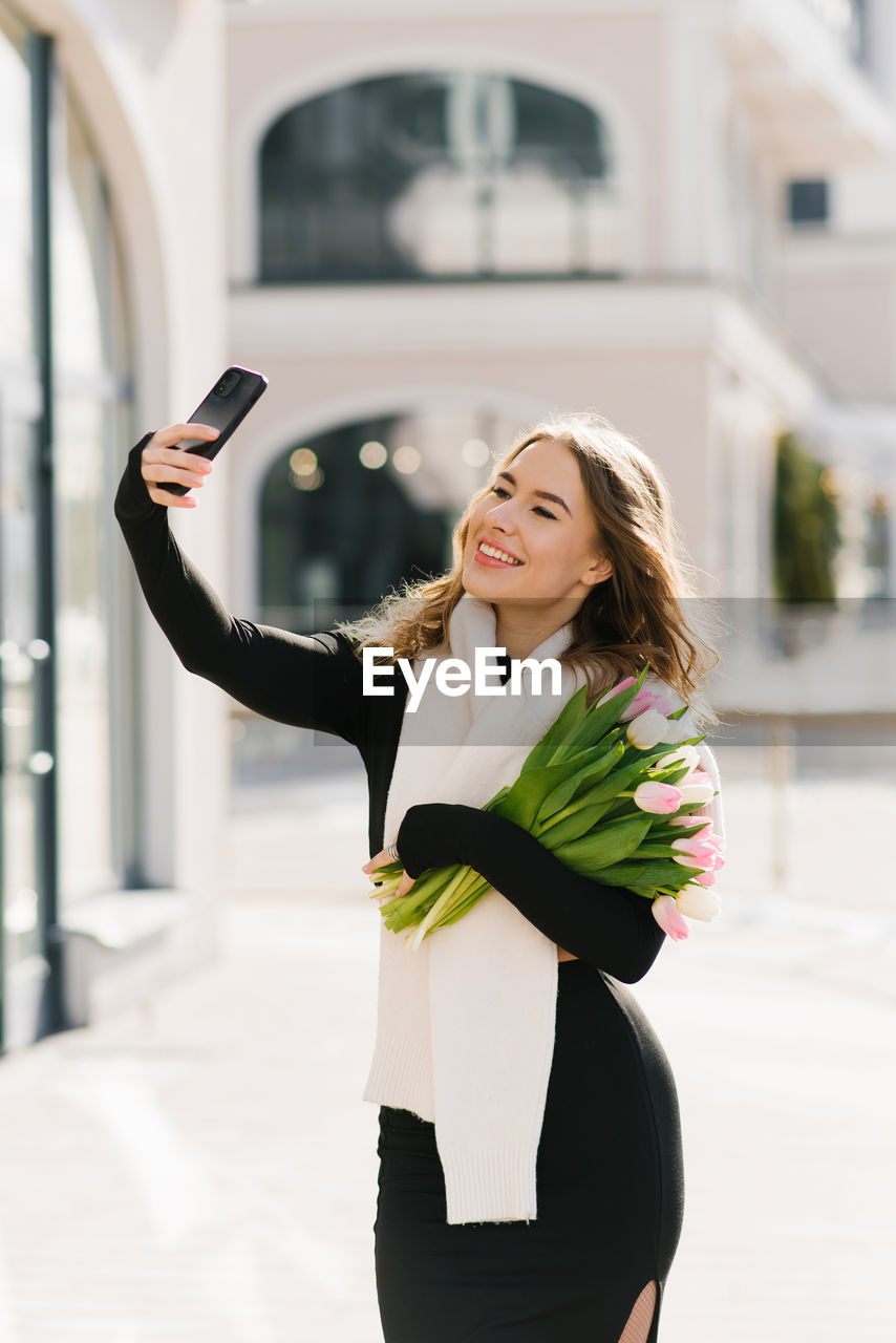 Happy young woman with a bouquet of tulips takes a selfie on her smartphone while walking 