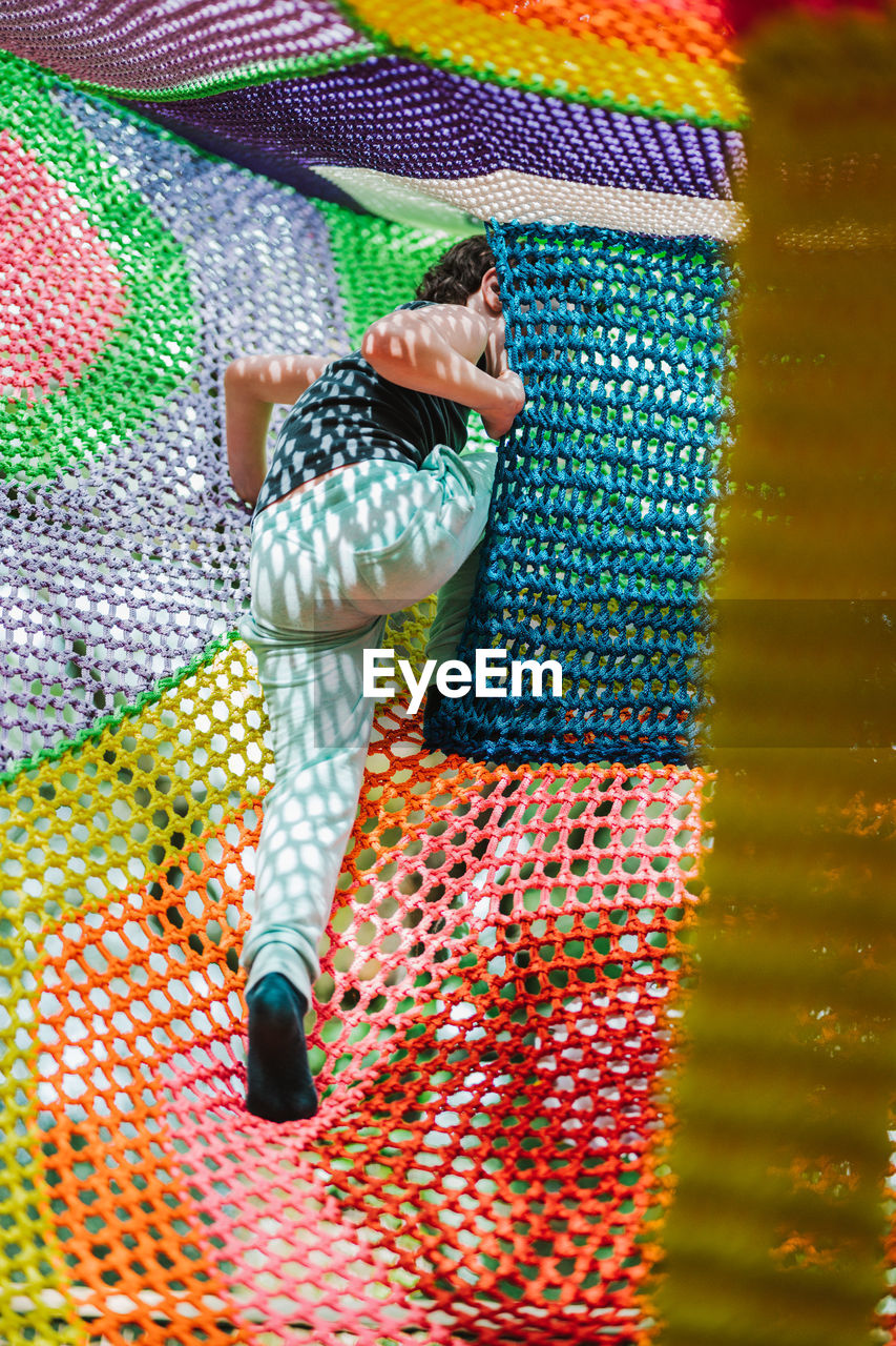 Adorable cheerful little boy with dark hair in casual clothes playing on colorful knitted trampoline in outdoor entertainment center