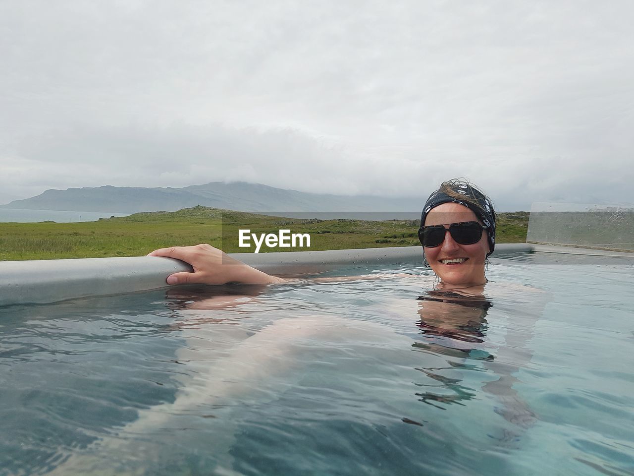Smiling woman in sunglasses swimming in pool against cloudy sky