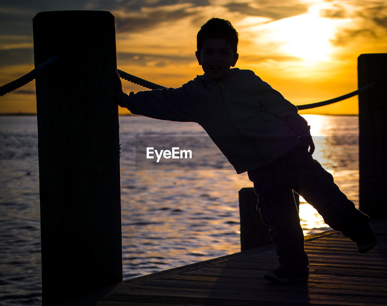 Silhouette boy standing on pier over sea against cloudy sky during sunset