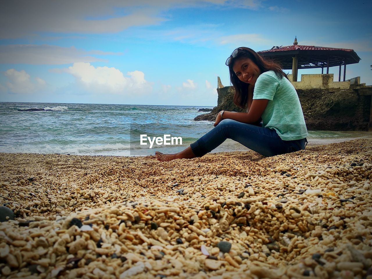 FULL LENGTH OF WOMAN SITTING ON ROCK AT BEACH AGAINST SKY