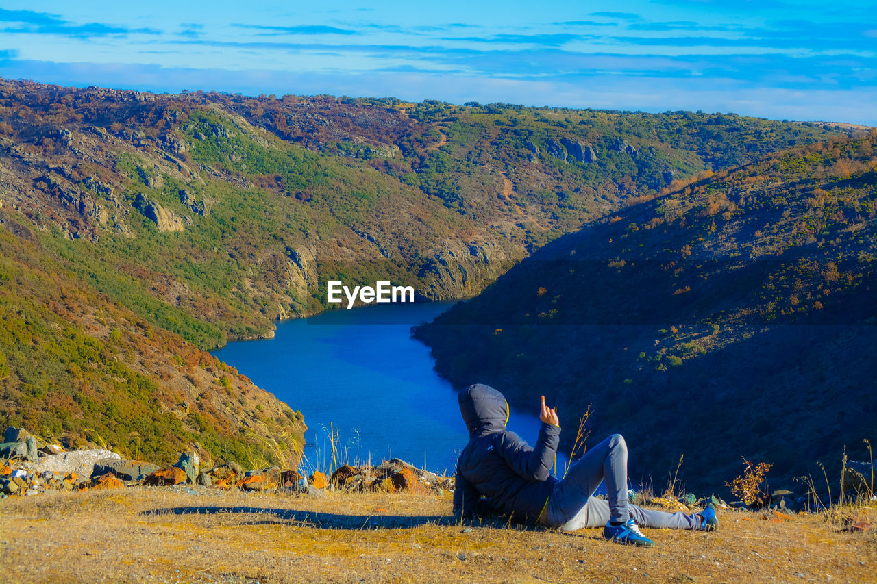 Man gesturing obscene gesture while lying on field over river against sky