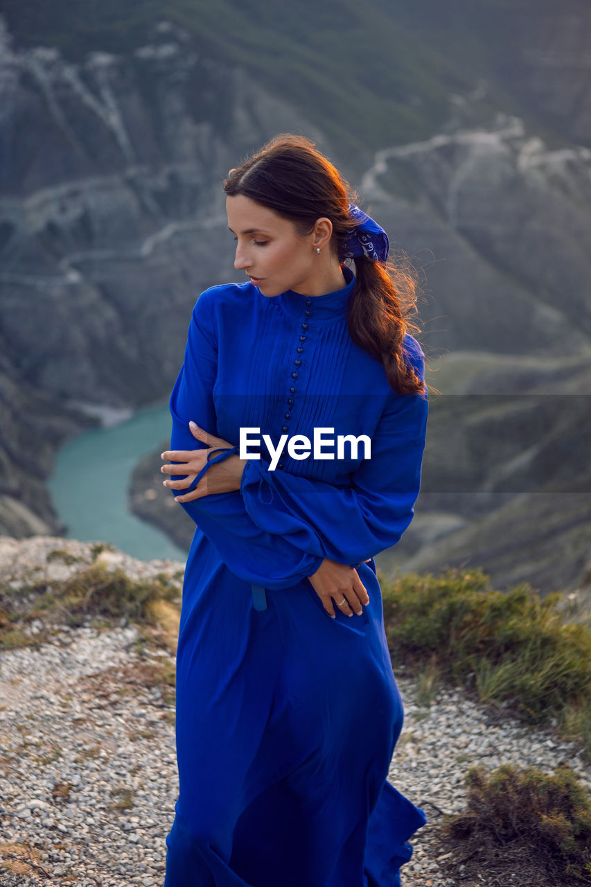 Brunette woman in a blue long dress stands on the edge of the sulak canyon in the evening at sunset