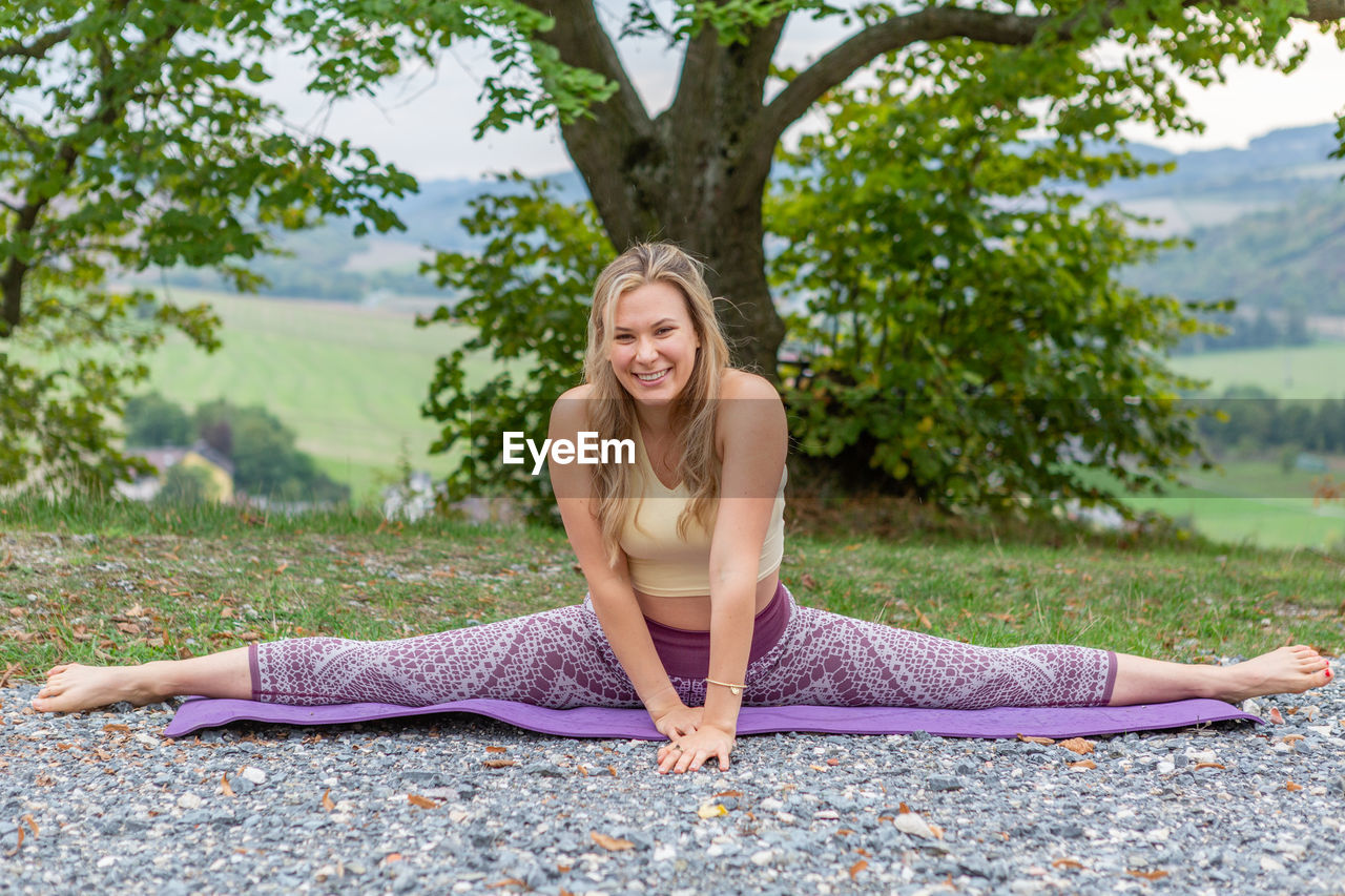 women, one person, adult, exercising, yoga, wellbeing, sitting, lifestyles, relaxation, tree, zen-like, plant, nature, meditating, sports, leisure activity, spirituality, clothing, full length, person, martial arts, stretching, young adult, smiling, physical fitness, tranquility, female, portrait, flexibility, blond hair, religion, long hair, balance, hairstyle, happiness, tranquil scene, cross-legged, outdoors, posture, day, vitality, exercise mat, sports clothing, lotus position, arm, human face, front view, beauty in nature, human leg, limb, summer, harmony, practicing, enjoyment, park, emotion, barefoot, brown hair