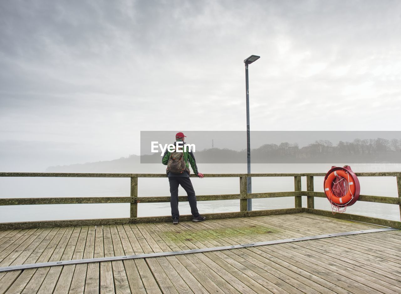 Rear view of man standing on pier over lake against sky