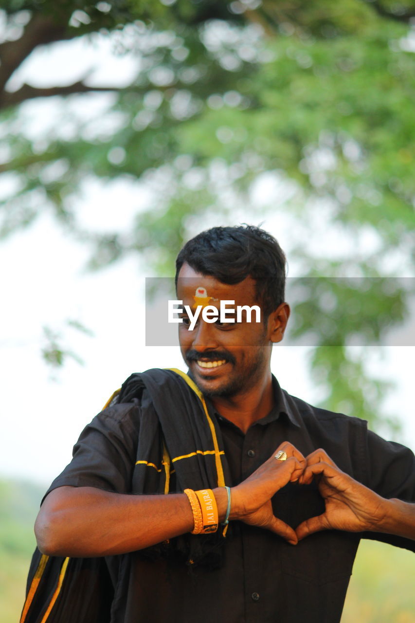 Man making heart shape standing against trees