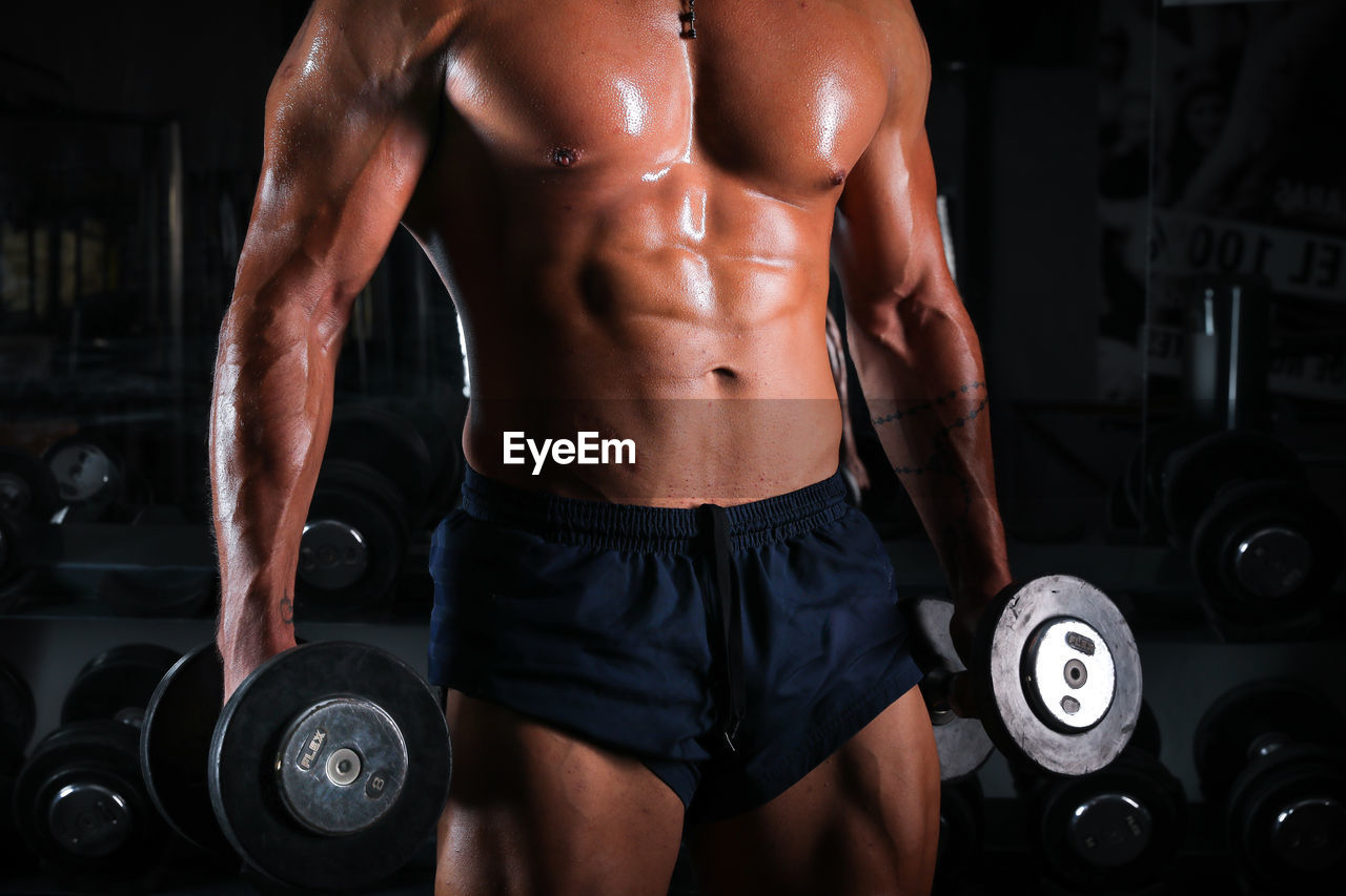 Close-up of shirtless muscular man lifting dumbbells at gym