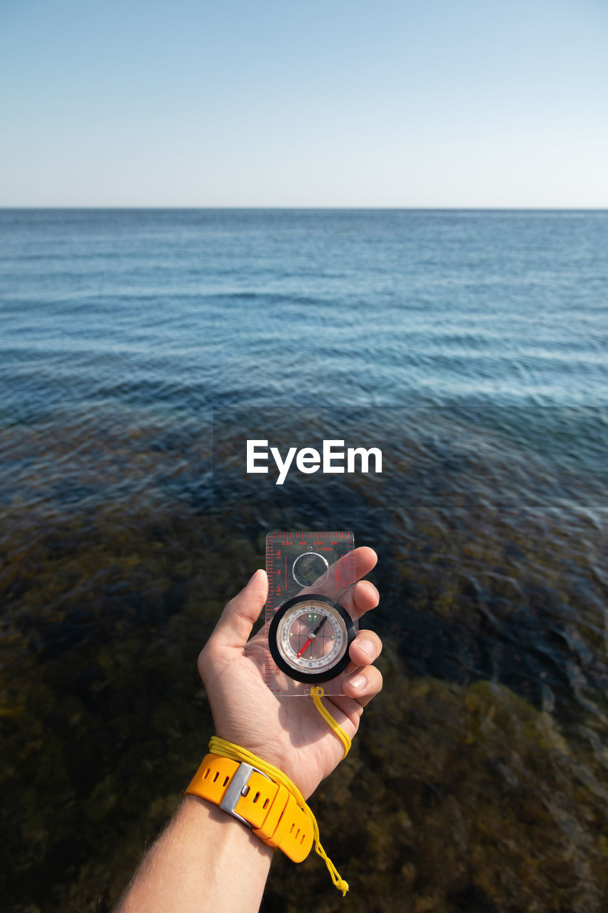 A man's hand with a wristwatch bracelet holds a magnetic compass against the background of the sea