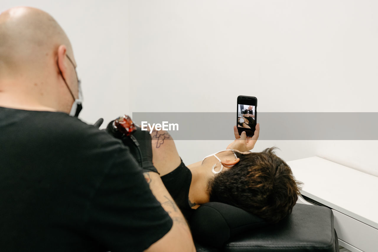 Content female in protective respirator lying on table and taking photo on smartphone while getting tattoo in salon with male master