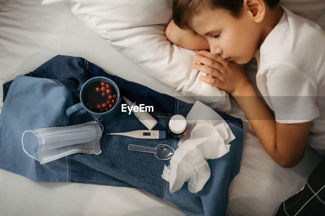 Sick child sleepping on bed. nearby there are tray with medicines, fruit drink, medical mask 