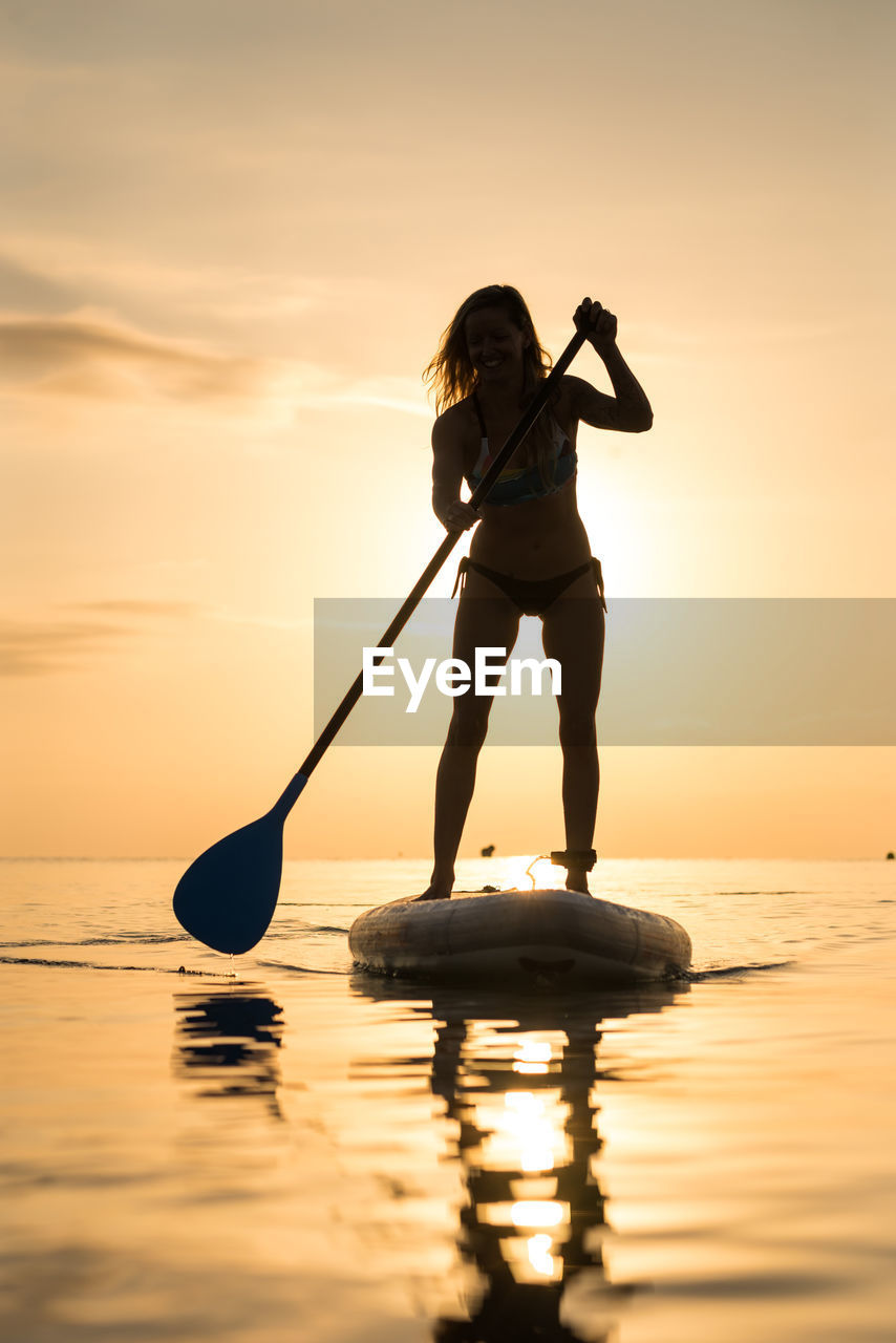 Back view of silhouette of unrecognizable female surfer standing on paddleboard and rowing against spectacular sun in sunset sky