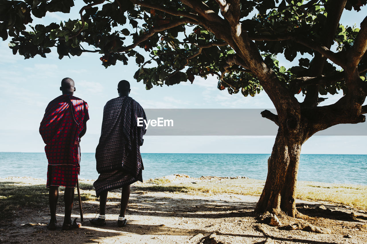 Two masai men in traditional clothes standing under big mkungu tree