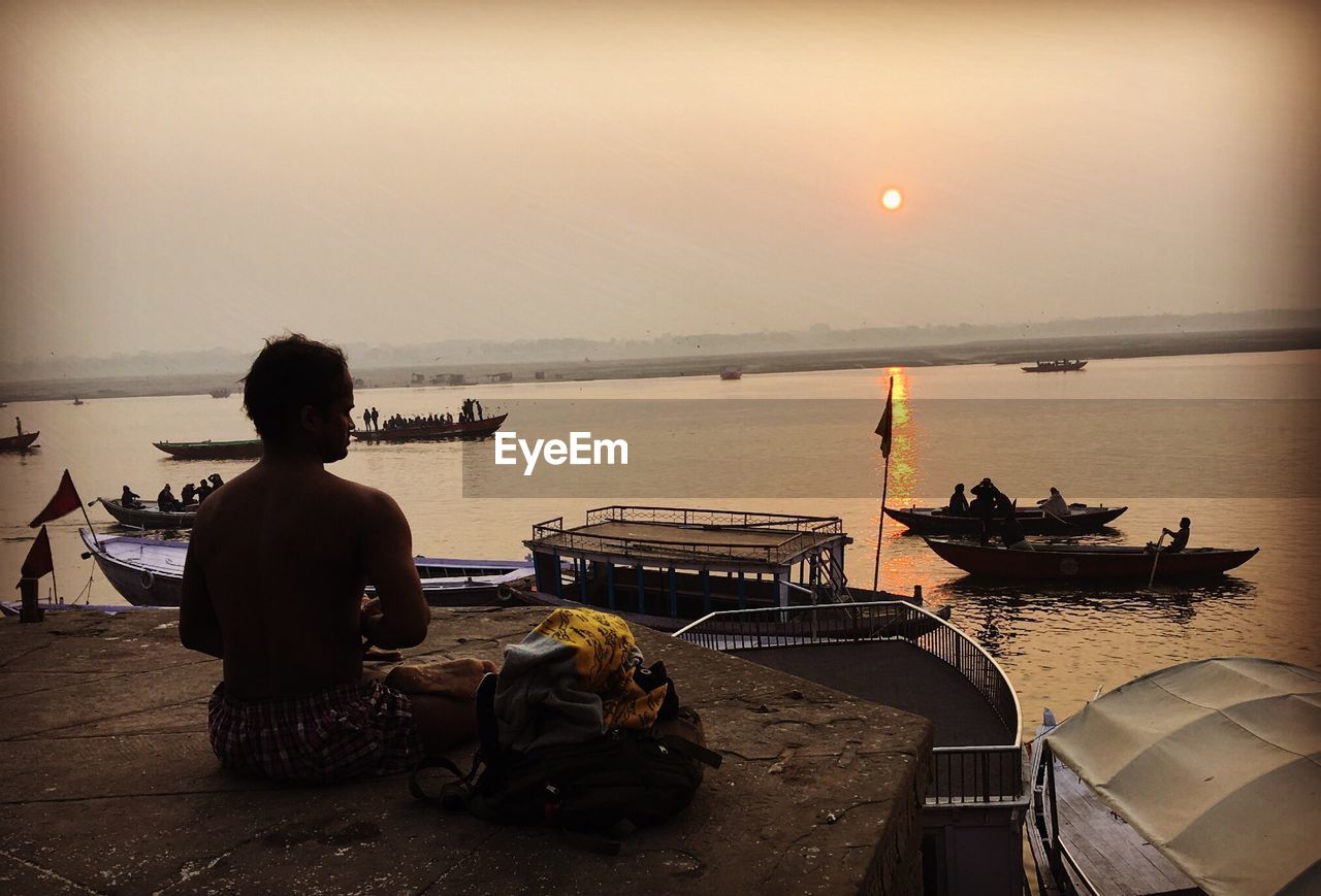 REAR VIEW OF MAN SITTING ON CHAIR BY SEA AGAINST SKY