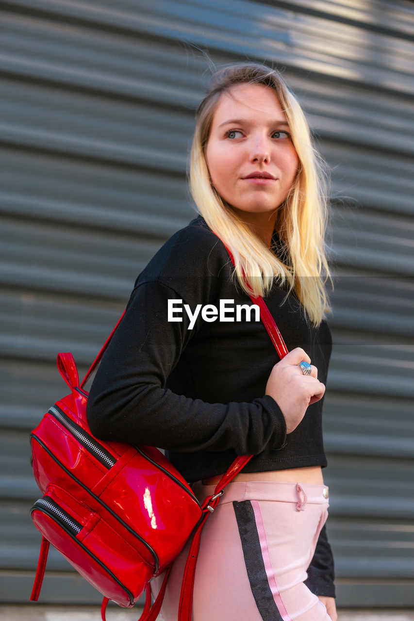 one person, women, adult, red, clothing, portrait, lifestyles, young adult, looking at camera, long hair, hairstyle, architecture, blond hair, three quarter length, person, human leg, female, bag, day, sitting, fashion, outdoors, smiling, luggage and bags, leisure activity, city, casual clothing, standing, handbag, happiness