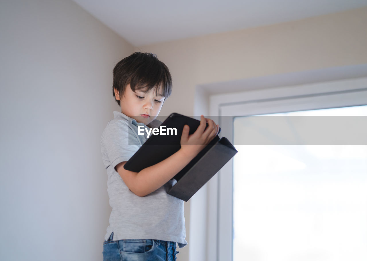 Boy holding mobile phone while standing against wall at home