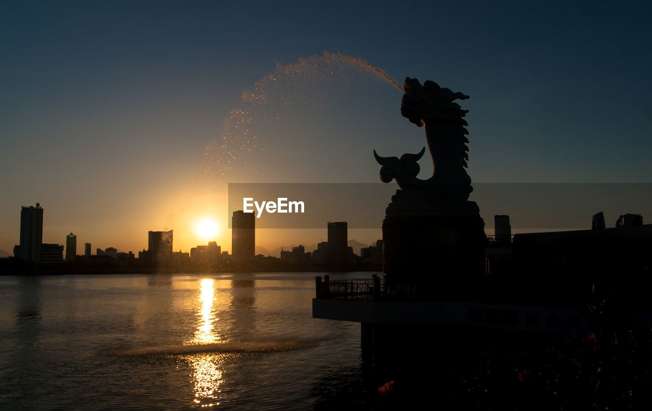 Silhouette carp dragon statue fountain by lake in city against sky during sunset