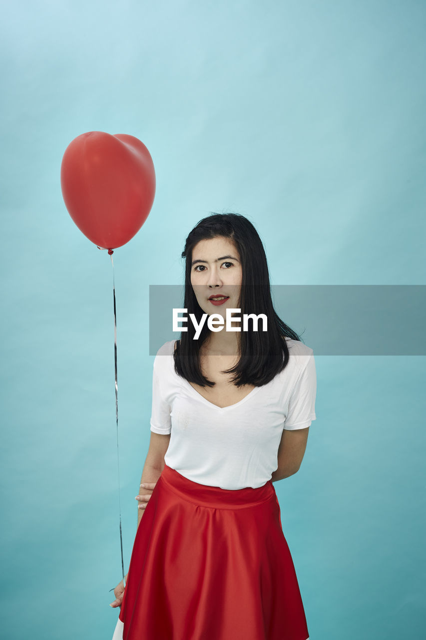 Portrait of woman holding heart shape balloon against blue background