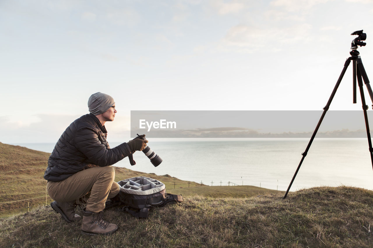 Side view of hiker looking away while holding slr camera on hill by sea against sky