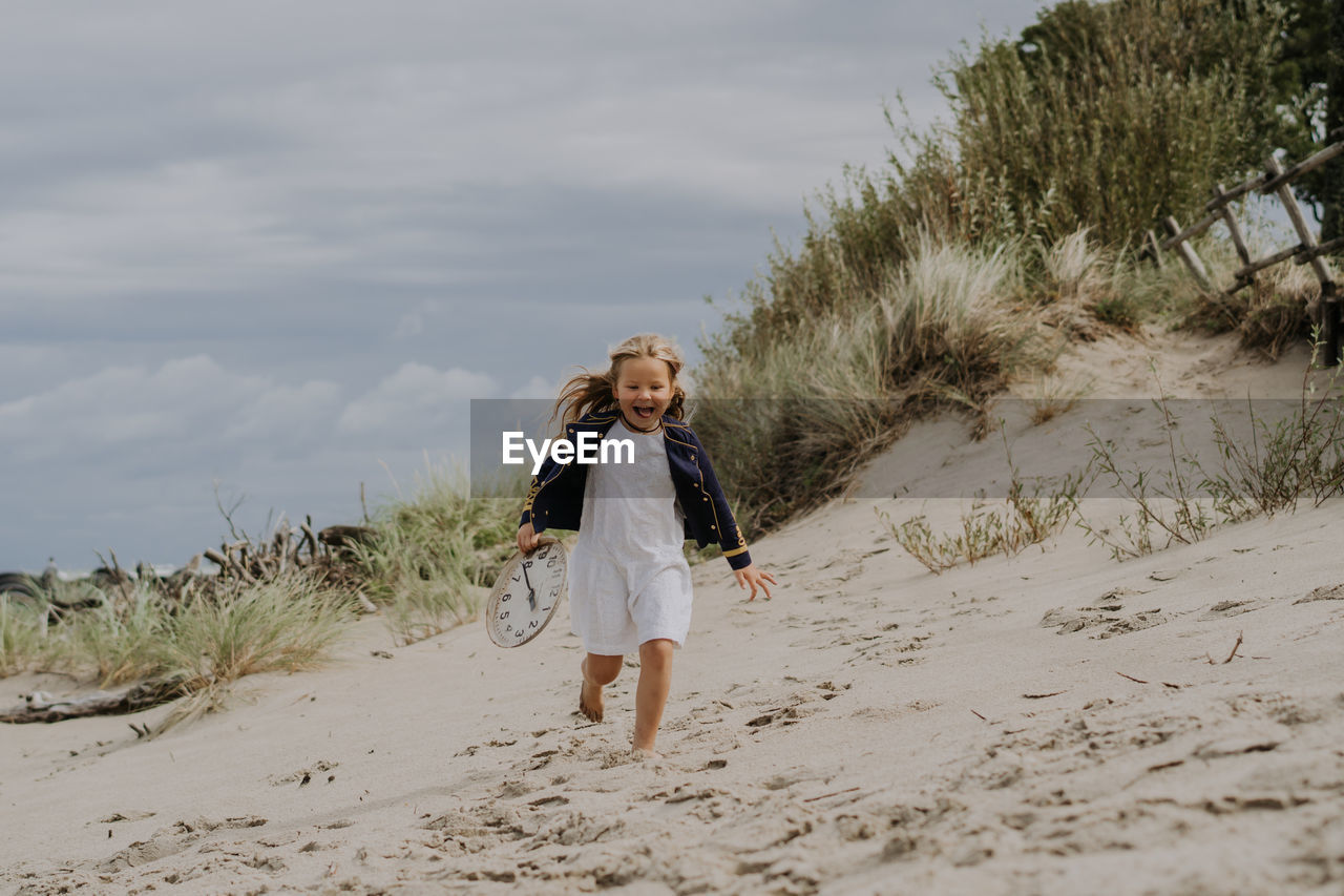 Child girl in jacket laughing and running with clock at the beach