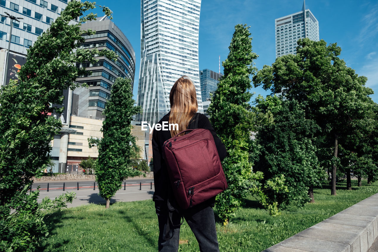 Back view of young woman with backpack standing alone on modern downtown high-rises skyscrapers