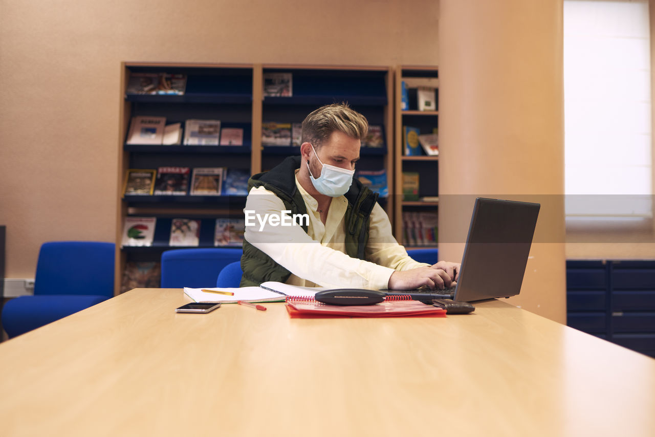 A young boy with a mask sitting and working with his laptop in the library