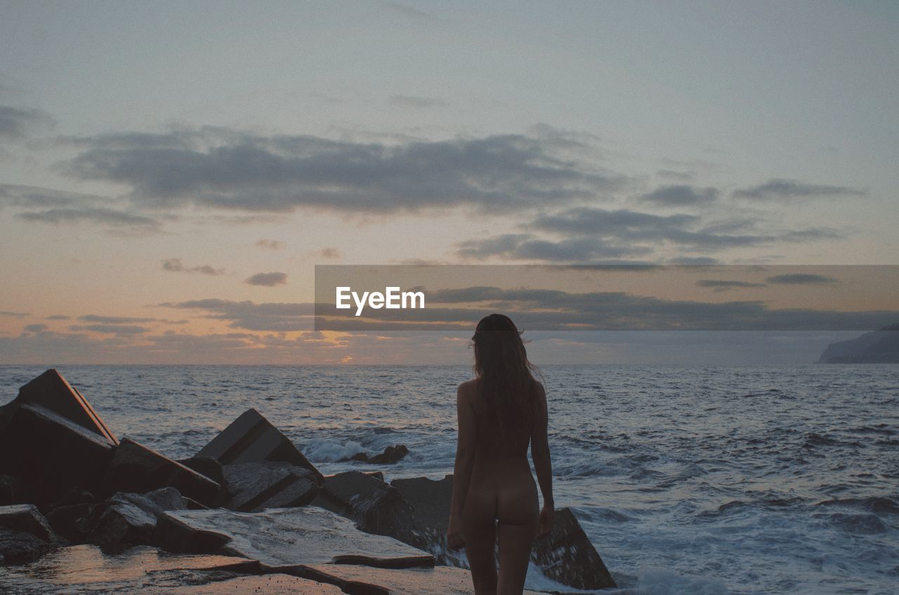 Naked woman standing at beach against sky during sunset