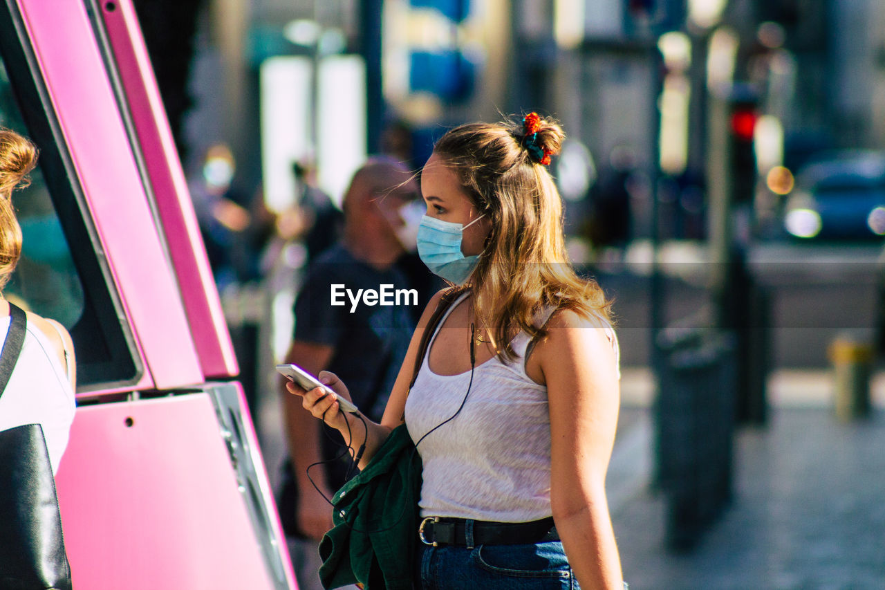 YOUNG WOMAN USING SMART PHONE WHILE STANDING ON CITY