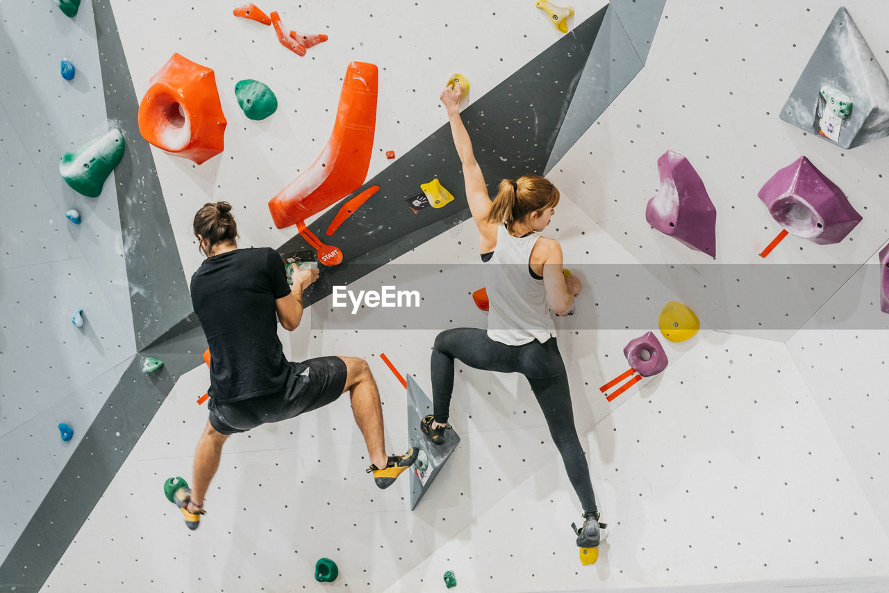 Back view of sportive couple in activewear climbing artificial wall with colorful grips during training together in light modern gym