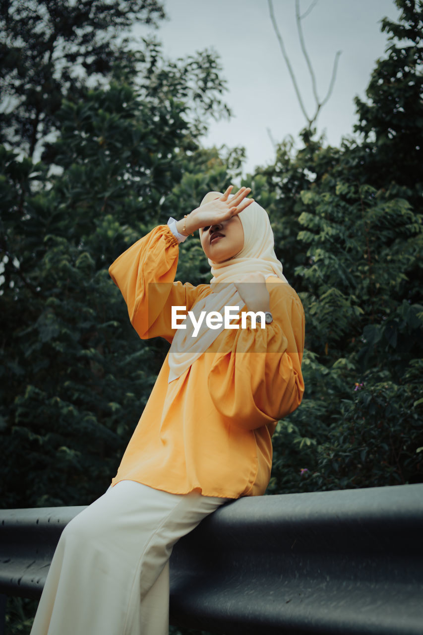 Woman wearing hijab sitting on railing against trees in forest