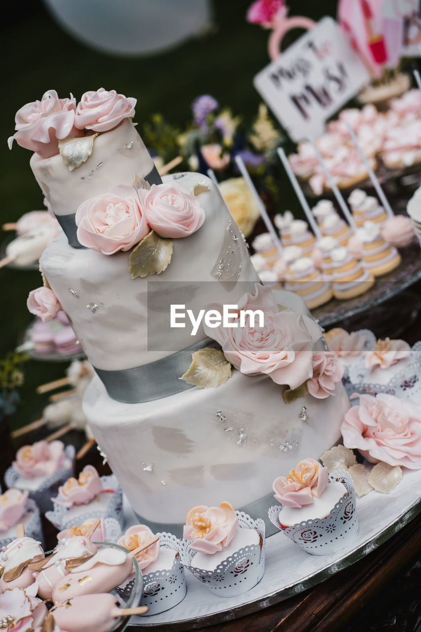 Big luxury white wedding cake with pink roses and cupcake decoration on the candy bar wooden table