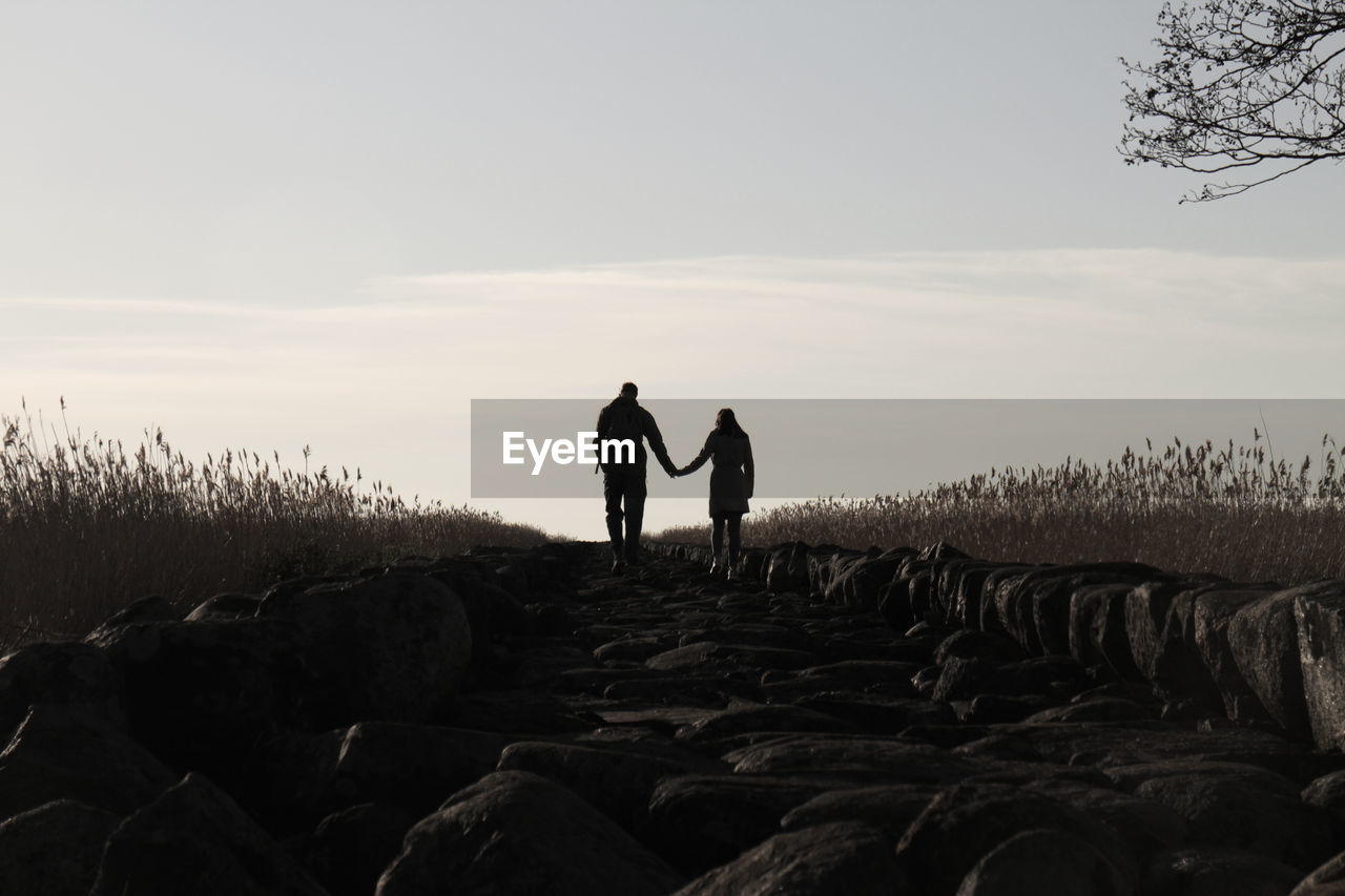 Rear view of silhouette couple holding hands while walking on rocks against sky during sunset