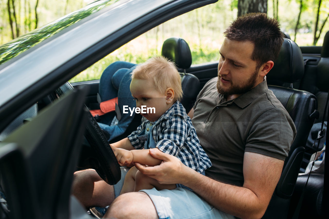 Dad shows his little son how to drive car while sitting behind wheel