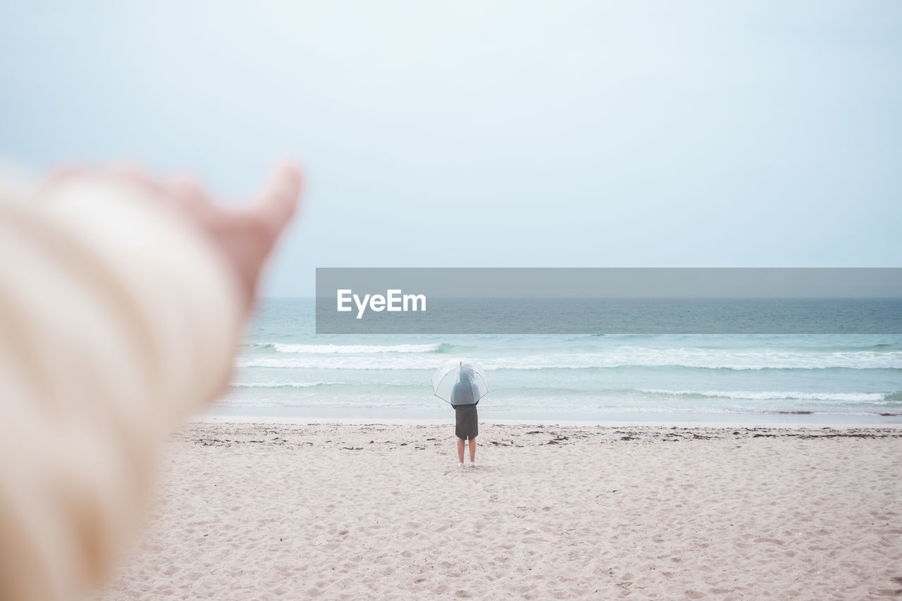 Crop anonymous person pointing finger away towards stormy ocean with lonely traveler with umbrella standing on sandy coast in overcast weather