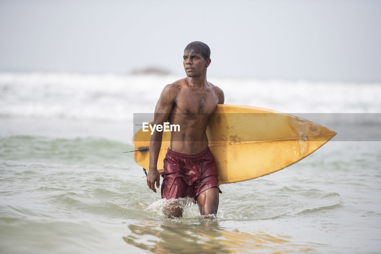 Shirtless man with surfboard wading in sea against sky