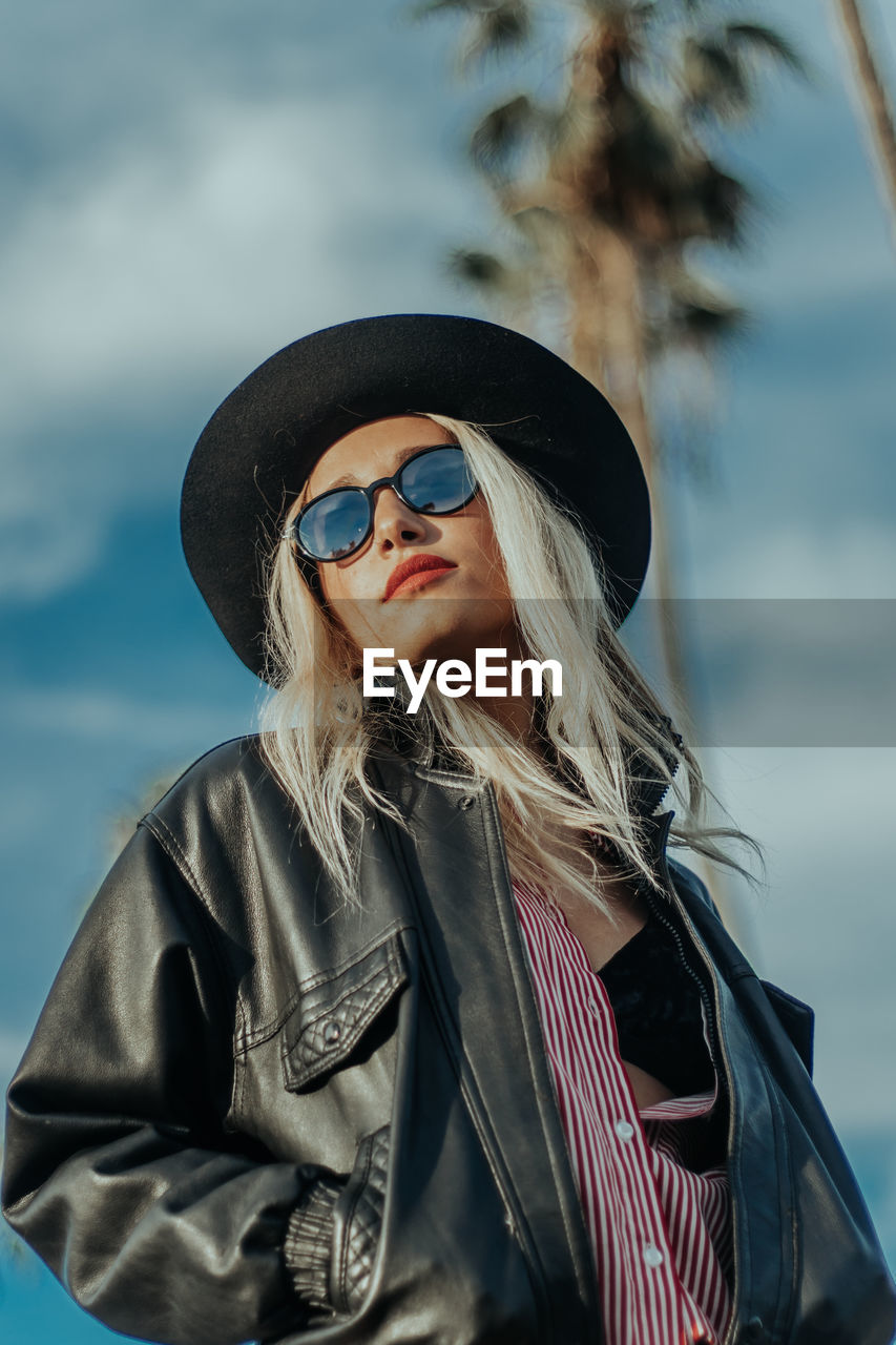 Low angle view of fashionable woman wearing sunglasses and jacket while standing against sky