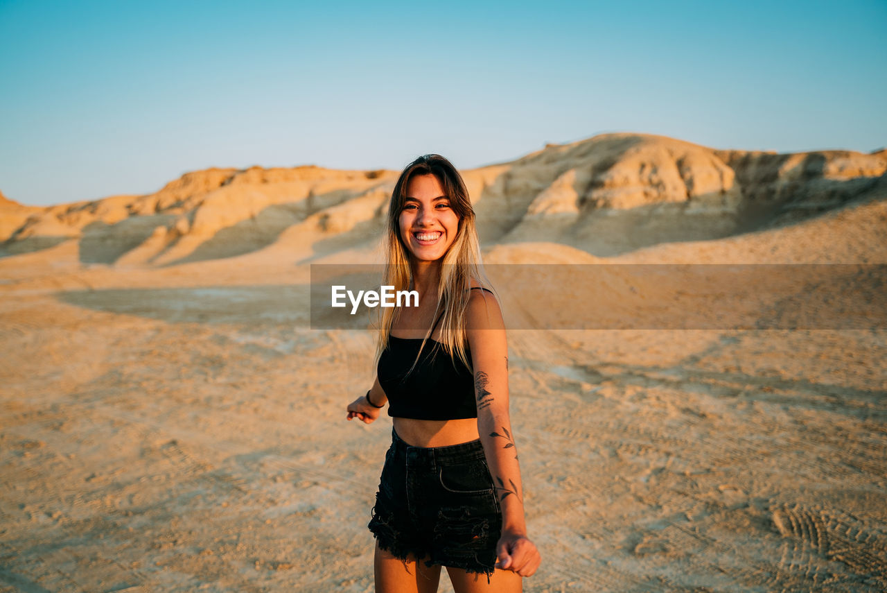 Positive female traveler standing in dry valley in bardenas reales and enjoying sunset in summer while looking at camera