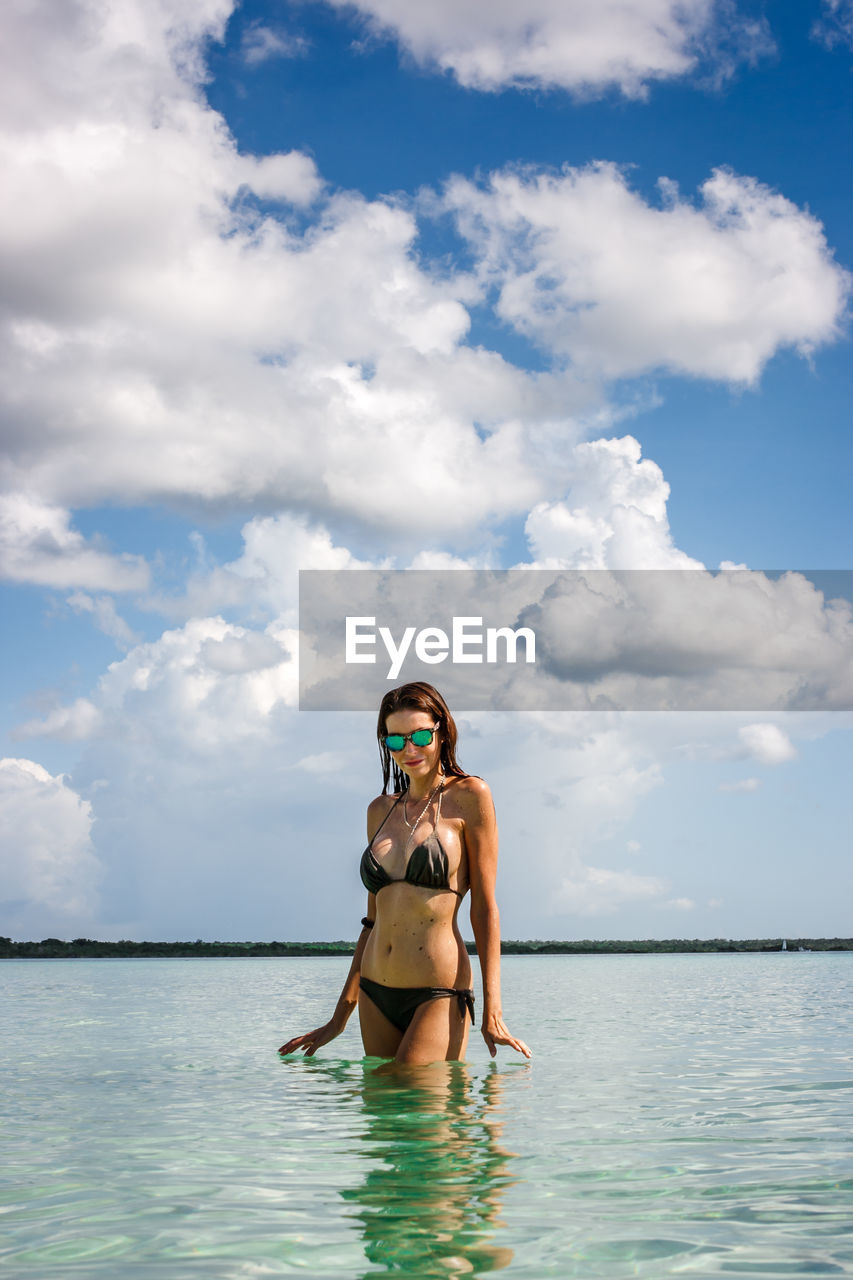 Young woman wearing sunglasses while standing in sea against cloudy sky
